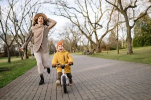 Happy mother running alongside her young child riding a balance bike in a park on a sunny day.